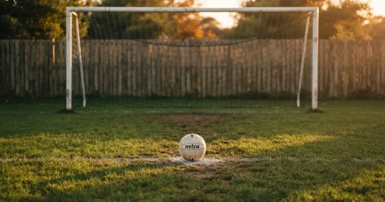 Ein Fußball liegt vor einem leeren Tor auf dem Rasen, das Netz hängt unbewegt, im Hintergrund ein verschwommener Zaun in warmem Abendlicht.