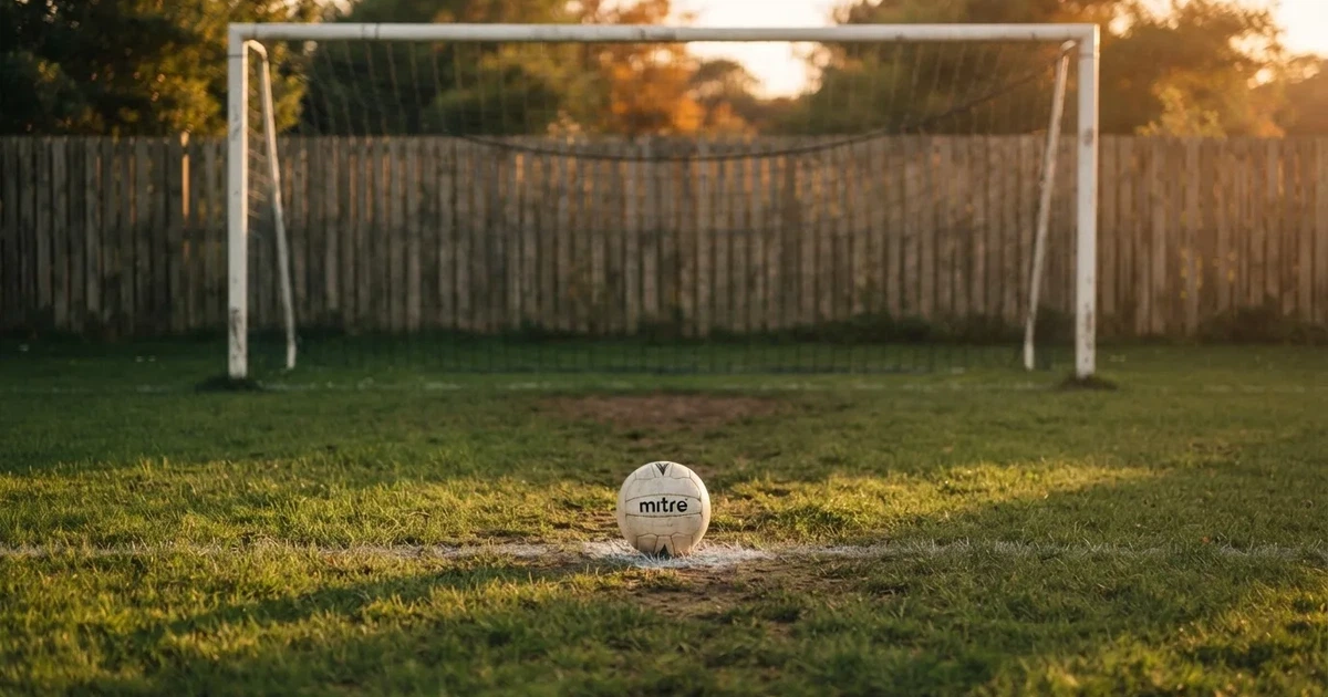 Ein Fußball liegt vor einem leeren Tor auf dem Rasen, das Netz hängt unbewegt, im Hintergrund ein verschwommener Zaun in warmem Abendlicht.