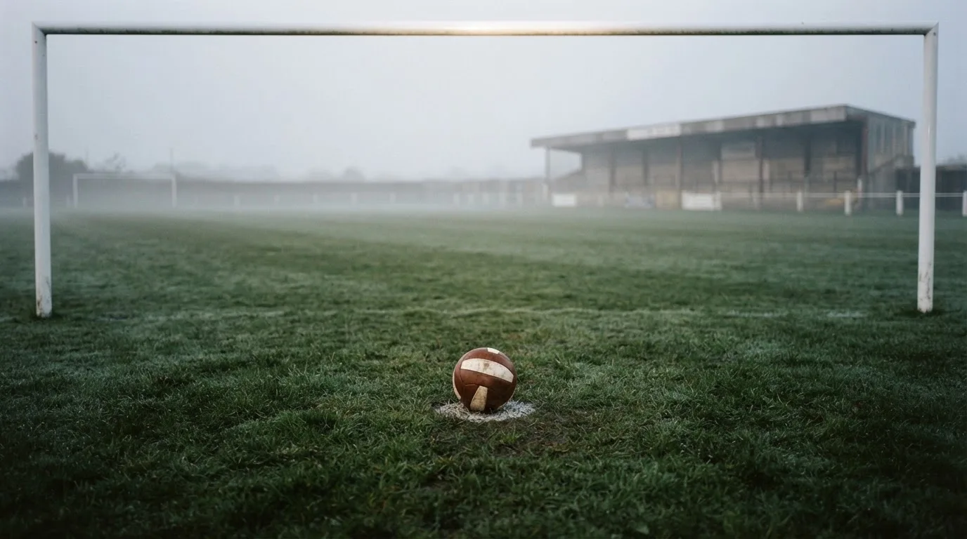 Ein Fußball liegt auf dem Anstoßpunkt eines leeren Rasenplatzes im Morgendunst, im Hintergrund eine ferne Tribüne.