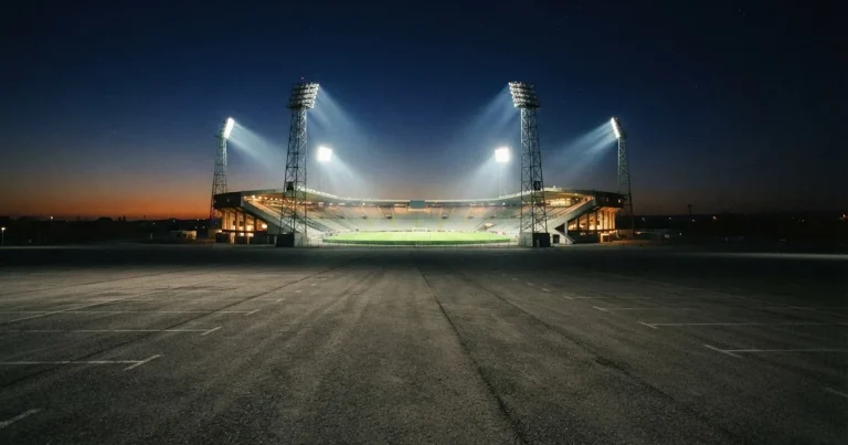 Ein Fußballstadion unter Flutlicht aus großer Entfernung gesehen, die Lichtkegel stehen im dunklen Abendhimmel über leeren Parkplätzen.