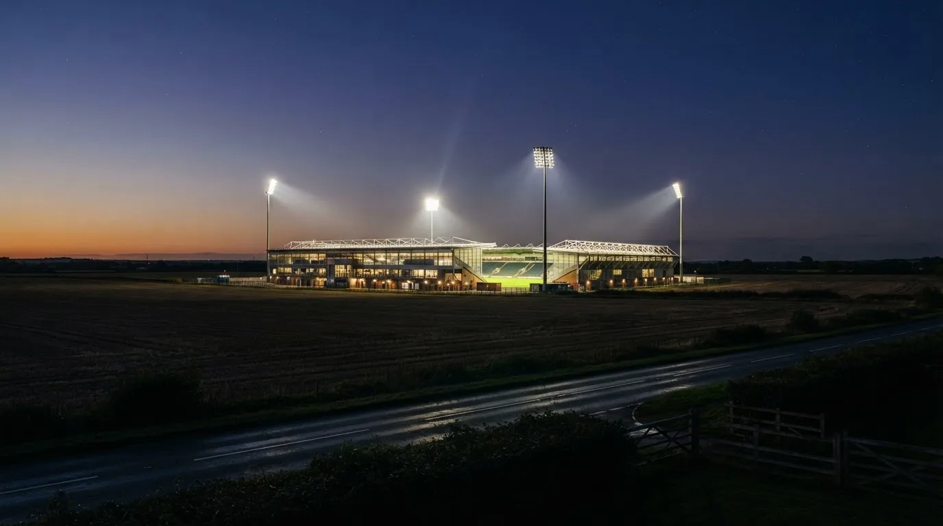 Ein Fußballstadion unter Flutlicht aus großer Entfernung gesehen, davor Felder und eine ruhige Straße in der Dämmerung.