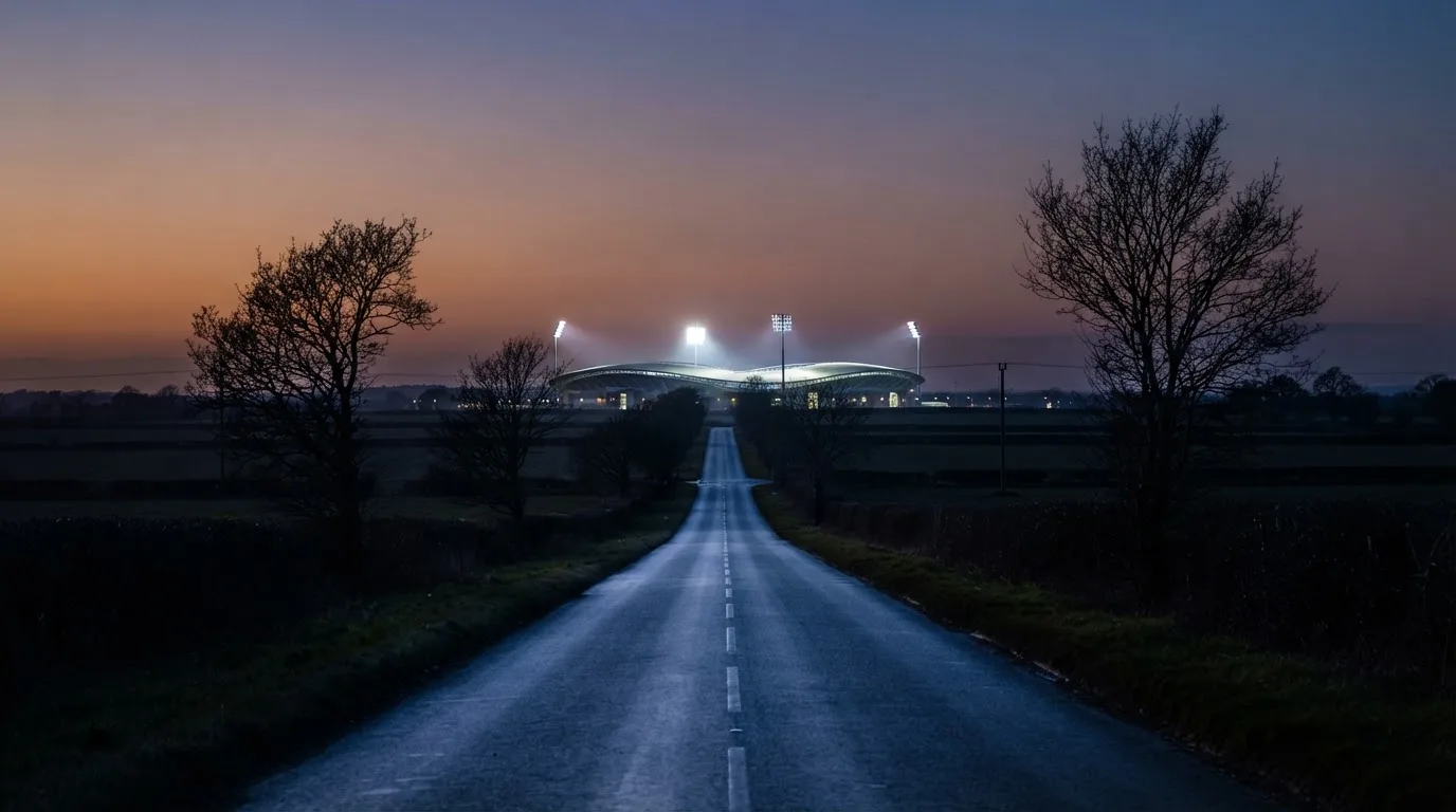 Ein Stadion unter Flutlicht aus großer Entfernung gesehen, davor eine dunkle Landstraße und einzelne Bäume in der Dämmerung.